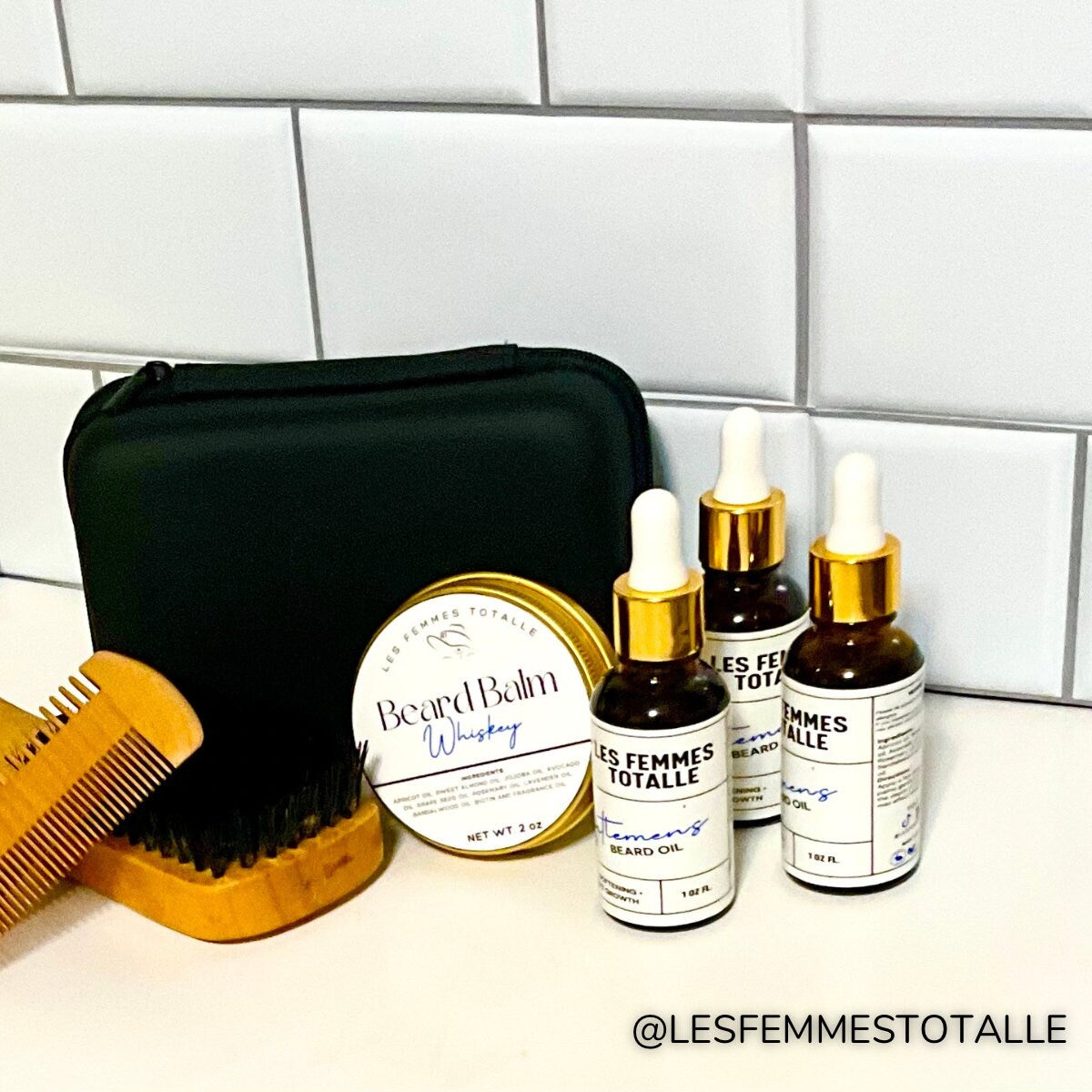 May include: A black zippered pouch, a wooden comb, a wooden brush, a jar of beard balm labeled &#39;Beard Balm Whiskey&#39; with a  logo, and three amber glass bottles of beard oil labeled &#39;Les Femmes Totalle&#39; with a  logo. The bottles have gold caps and droppers. The products are arranged on a white tiled surface.